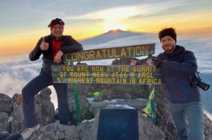 Zwei Bergsteiger stehen vor dem Gipfelschild des Mount Meru, dem Socialist Peak